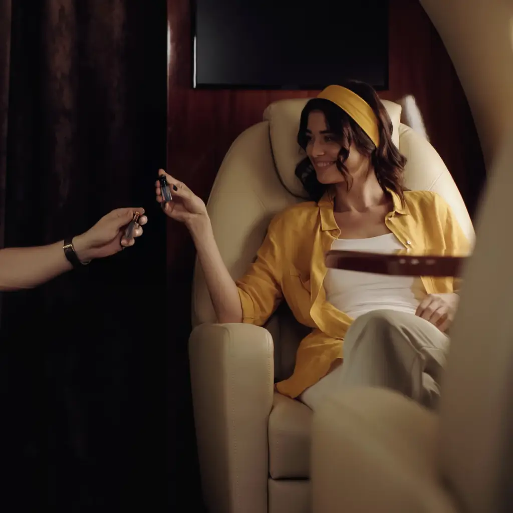 a smily beautiful young women wearing white clothing with yellowish jacket, with big hair band in same colour of the jacket, while she is travelling in a luxurious private flight, she is carrying a small sample of oil fragrance that she was showing to the side person (male hand) who holds a similar bottle of fragrance bottle - AeroNova Flight Support